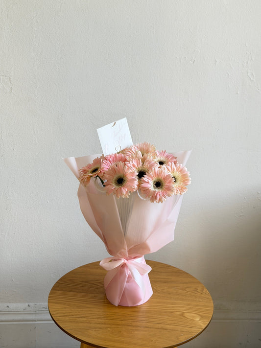 Ramo Dinorah de gerberas rosa claro con papel coreano y listón rosa, sobre una mesa de madera.